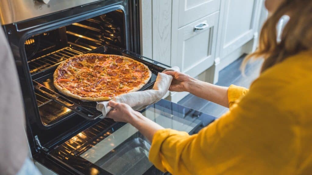 Person wearing a yellow shirt uses a towel to remove a baked pizza from an open oven in a modern kitchen.