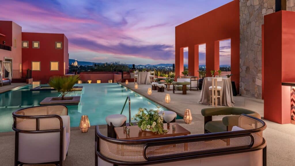 Outdoor lounge with modern seating, pool, and lanterns at sunset; vibrant pink and purple sky over red and stone buildings.