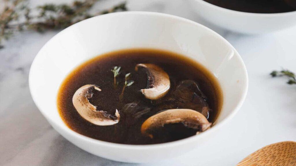 A white bowl filled with dark broth, containing three mushroom slices and a small sprig of thyme.