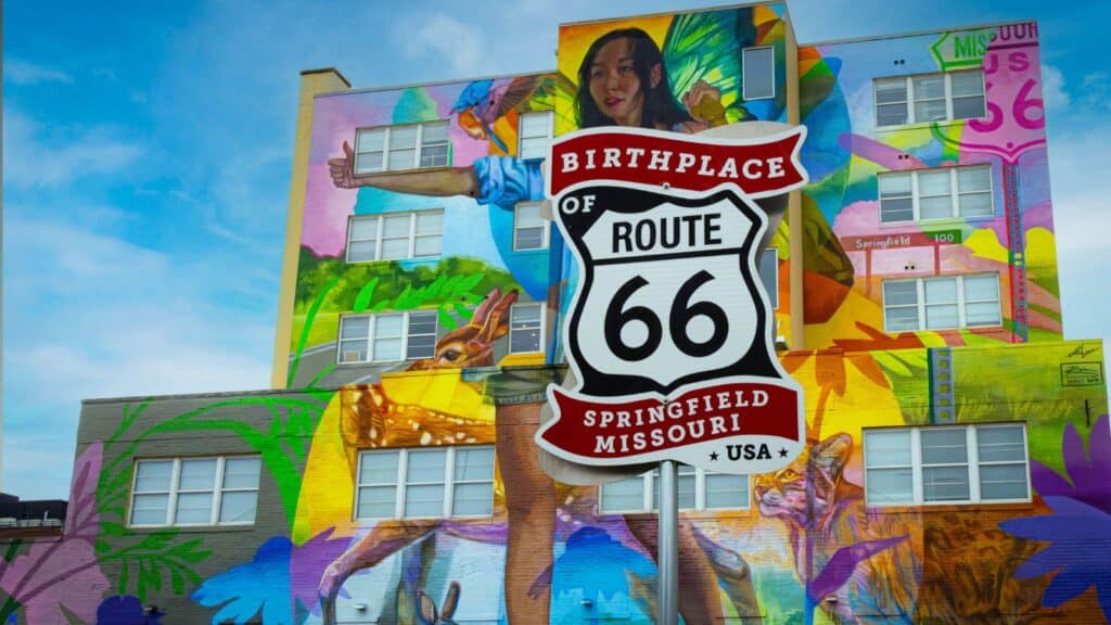 A Route 66 birthplace sign stands in front of a colorful mural on a building in Springfield, Missouri, celebrating Route 66 turns 100 with a hitchhiking woman and vibrant elements.
