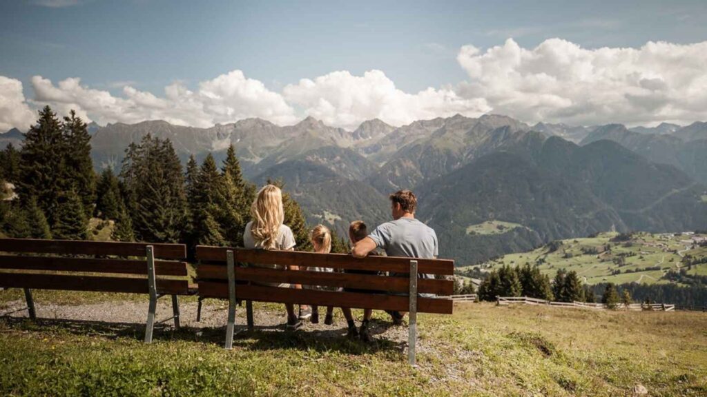 A family of four sits on a bench outdoors, dreaming of european trips as they face distant mountains and a green valley under a partly cloudy sky.