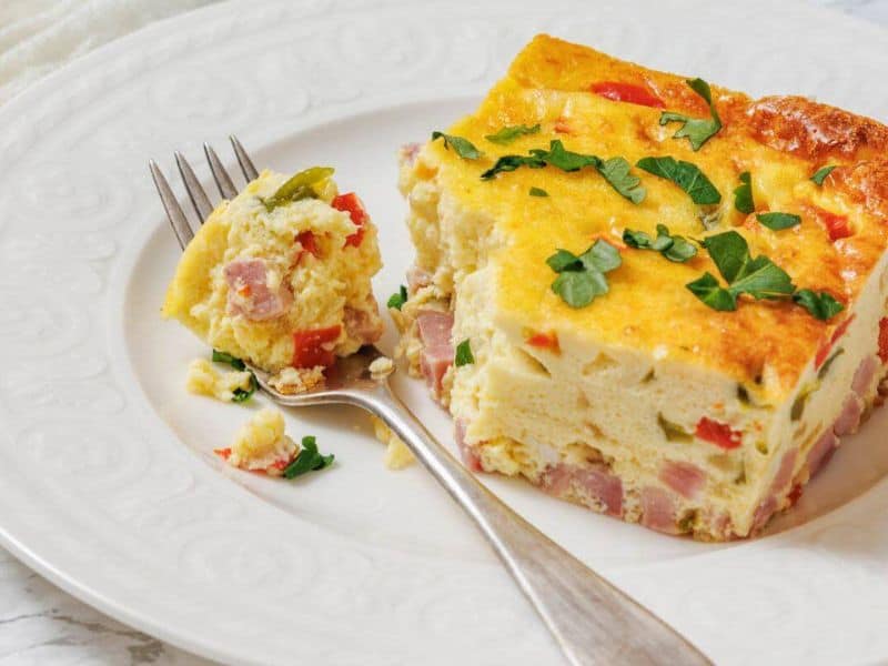 A slice of baked Western omelet with ham, red peppers, and herbs is served on a white plate with a fork holding a bite-sized piece.