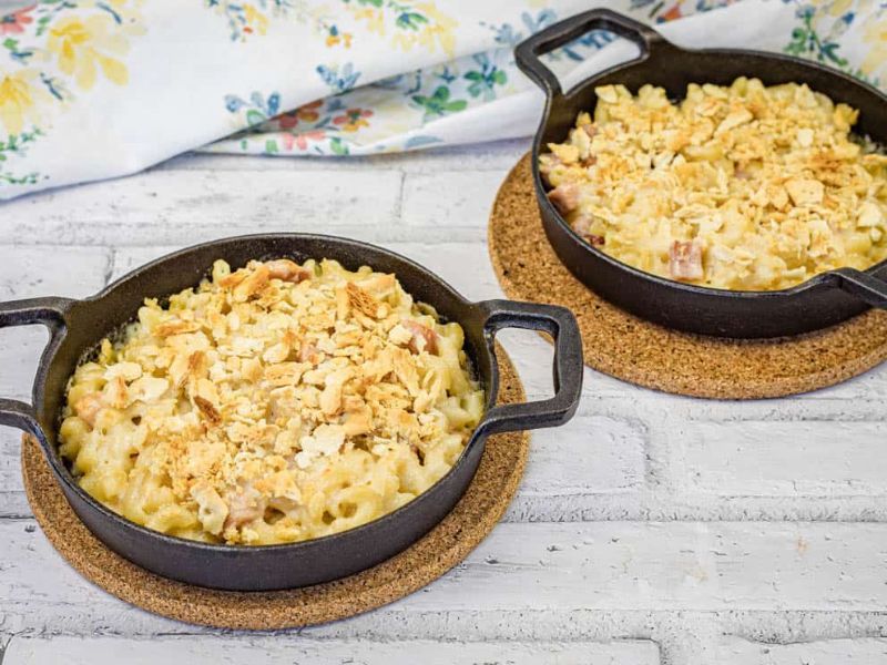 Two black cast iron pans filled with mac and cheese sit on cork trivets on a white wooden surface.