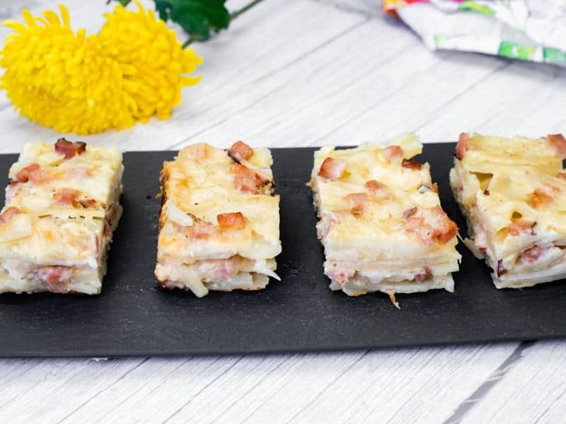 Four slices of Scalloped Potatoes with Ham and Cheese on a black slate board, placed on a light wooden surface.