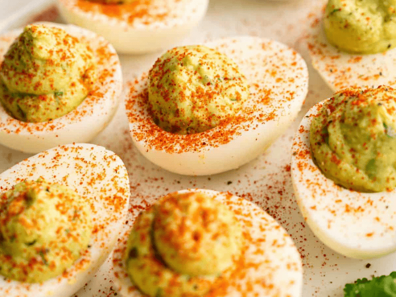 A close-up of deviled eggs with a creamy green filling, topped with a sprinkle of red paprika, arranged on a white plate.