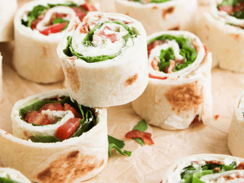Several tortilla pinwheels filled with leafy greens, tomato, and possibly cheese or meat, arranged on parchment paper.