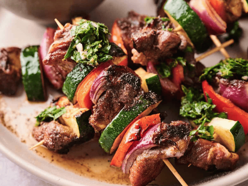 Grilled meat and vegetable skewers with zucchini, red onion, and red bell pepper, garnished with chopped herbs, served on a beige plate.