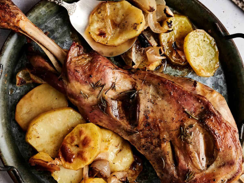 Roasted lamb leg on a platter with sliced potatoes, lemon, and herbs. A knife is placed beside the dish.