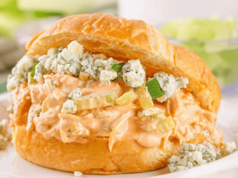A croissant sandwich filled with buffalo chicken and blue cheese, served on a white plate with celery sticks in the background.