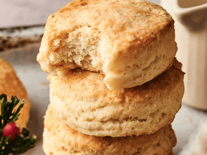 Three golden-brown biscuits are stacked, with the top one missing a bite. A small sprig of greenery with red berries is visible in the corner.