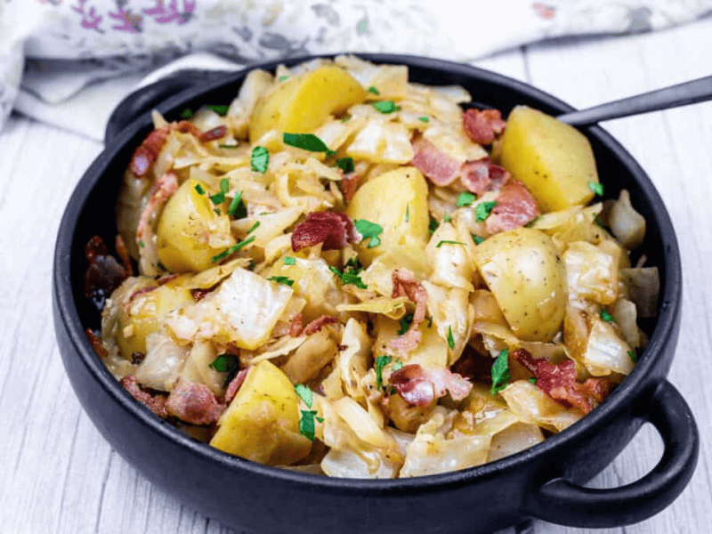 A black bowl filled with Cabbage & Potatoes with Bacon with a spoon on the side.