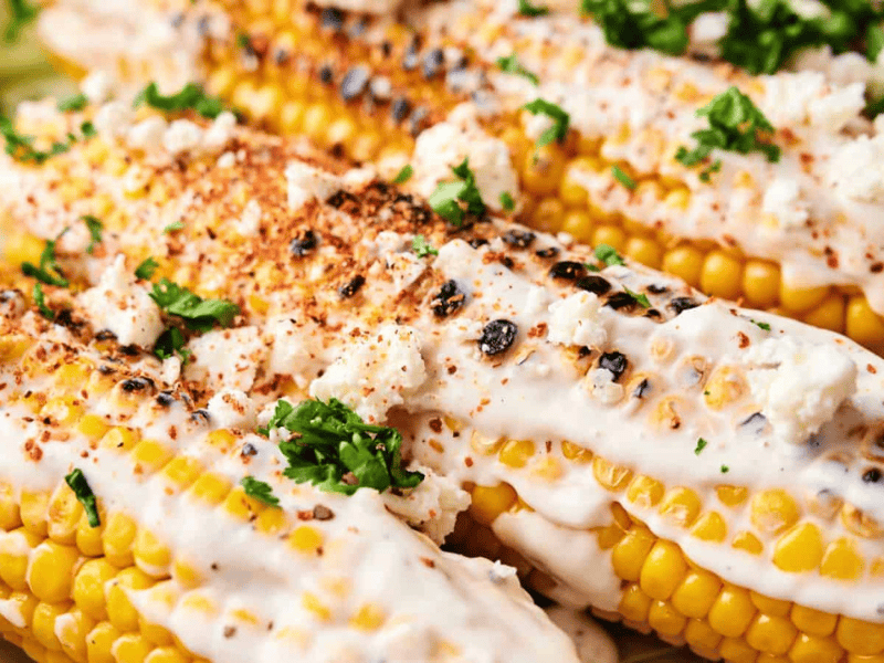 Close-up of Mexican Street Corn on the cob topped with creamy sauce, crumbled cheese, chili powder, and chopped cilantro.