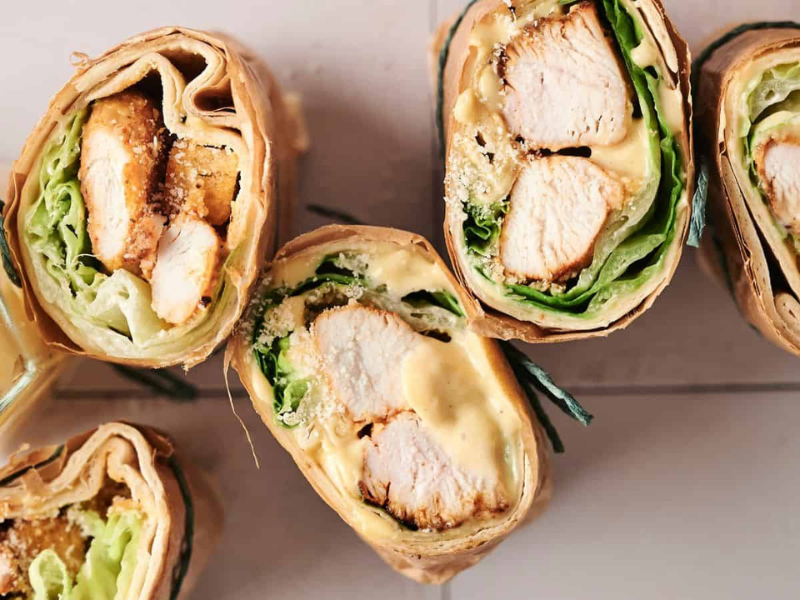 Close-up of sliced chicken wraps with lettuce, cheese, and a creamy sauce, next to an open jar of mustard on a wooden surface.