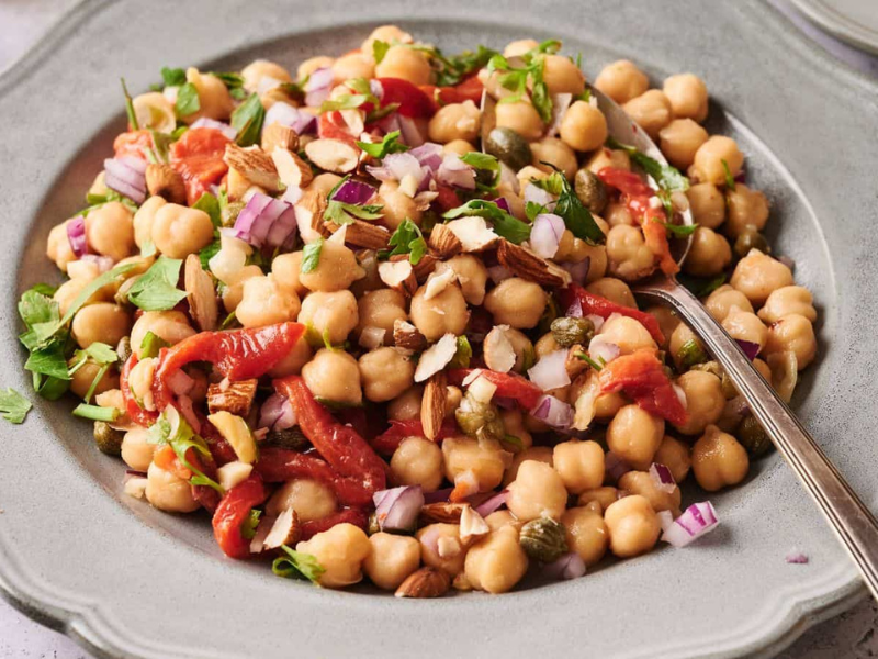 A plate of chickpea salad with chopped red onions, roasted red peppers, herbs, and sliced almonds, served with a spoon.