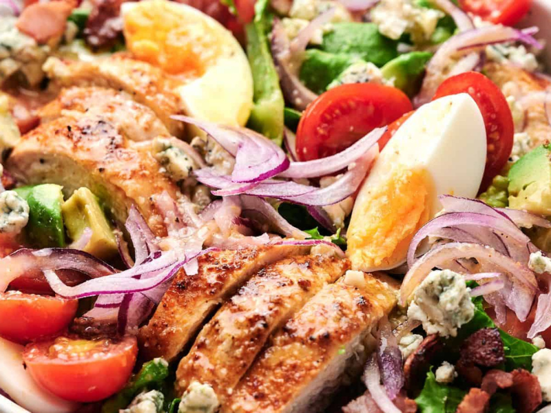 A close-up of a Cobb salad featuring sliced chicken, cherry tomatoes, avocado, boiled eggs, red onion, bacon bits, and blue cheese crumbles on a bed of lettuce.