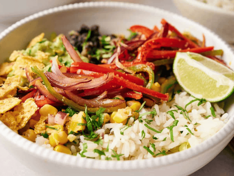 A Copycat Chipotle burrito bowl, filled with rice, fajita veggies, corn salsa, tortilla chips. and topped with a lime wedge.