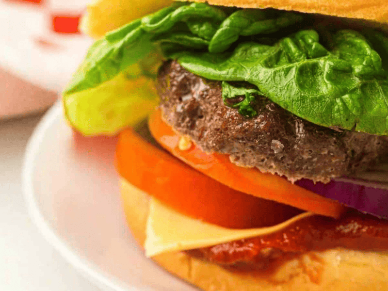 A close-up of a hamburger with lettuce, tomato, onion, cheese, ketchup, and a beef patty in a bun, served on a white plate.