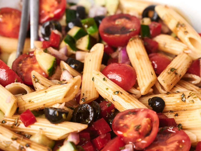 A bowl of vibrant pasta salad with penne, cherry tomatoes, black olives, diced cucumbers, red onions, and a drizzle of dressing.