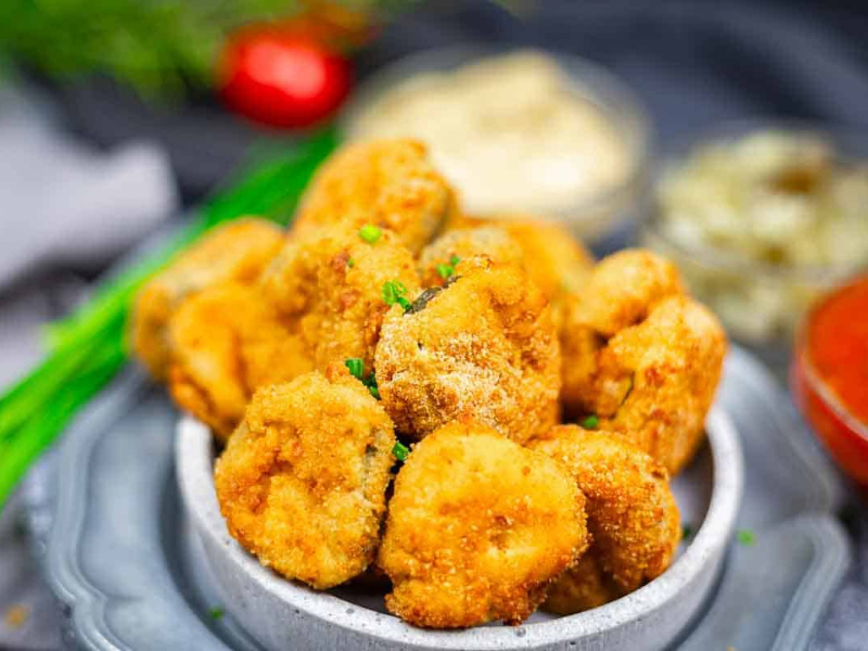 A bowl filled with breaded and fried pickle bites, garnished with chopped herbs, with dipping sauces and blurred vegetables in the background.