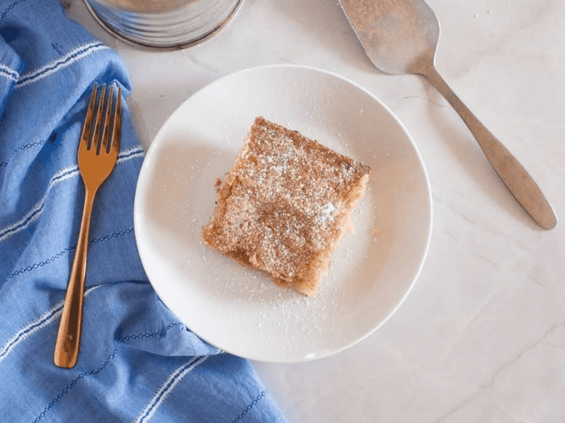 A square piece of Gooey Butter Cake. topped with powdered sugar on a white plate, next to a gold fork, a blue cloth, and a metal cake server on a marble surface.
