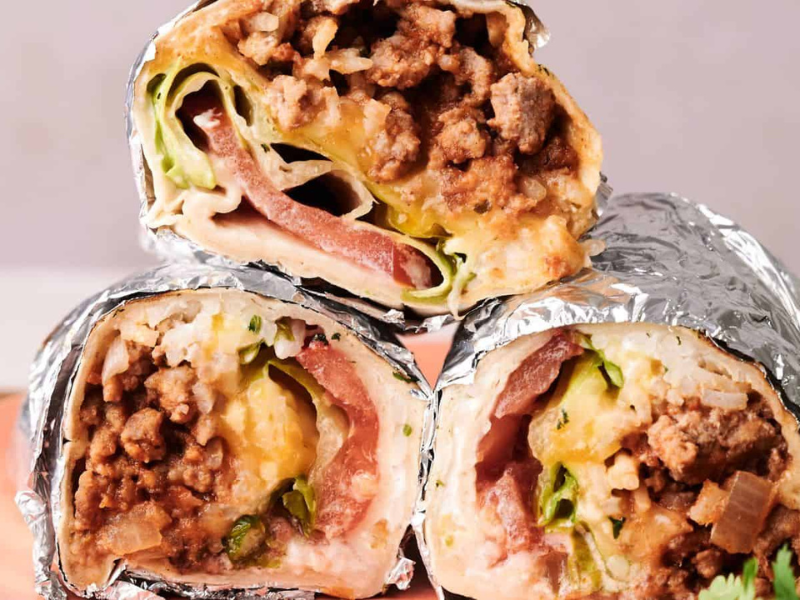 Three foil-wrapped burrito halves filled with ground meat, cheese, lettuce, and tomato are stacked on a pink plate.