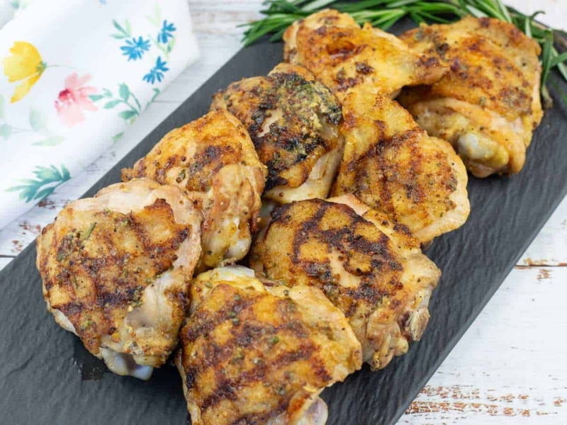Grilled Dijon rosemary chicken thighs are arranged on a rectangular black slate platter, garnished with fresh rosemary, next to a white floral napkin on a wooden surface.