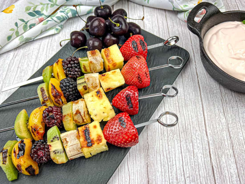 Four pieces of grilled fruit skewers on a slate plate with a bowl of cherry dip.
