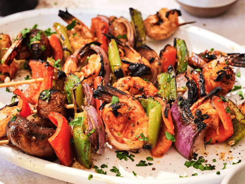 A white plate holds grilled shrimp and vegetable skewers with mushrooms, bell peppers, onions, and tomatoes, garnished with chopped herbs.