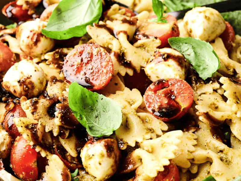 Bowtie pasta salad with cherry tomatoes, mozzarella balls, fresh basil leaves, and a drizzle of balsamic glaze in a green bowl.