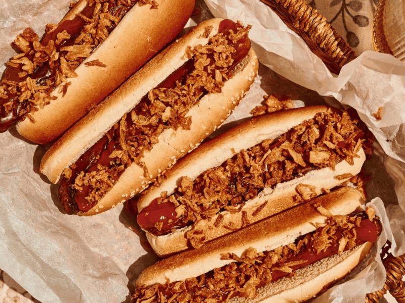 Four hot dogs in buns topped with crispy fried onions are arranged on parchment paper in a basket.