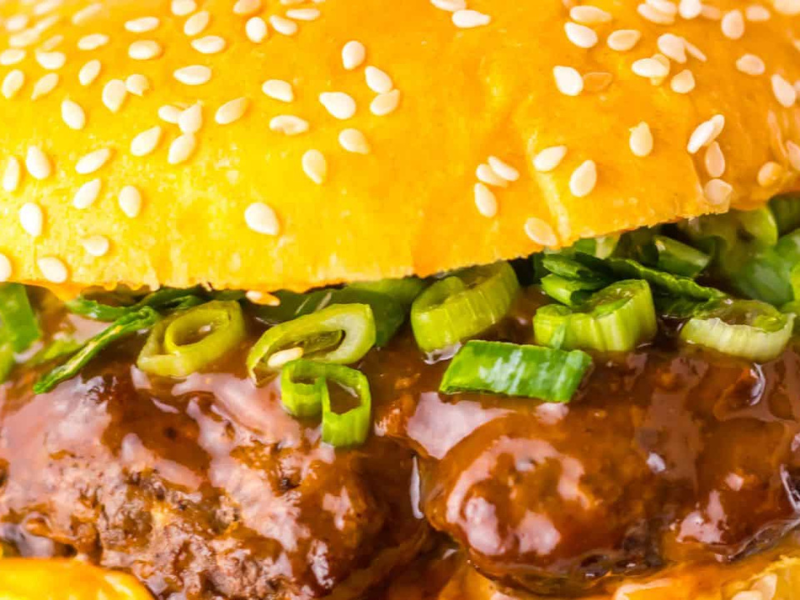 Close-up of a hamburger with a sesame seed bun, beef patty, green onions, and rich brown sauce.