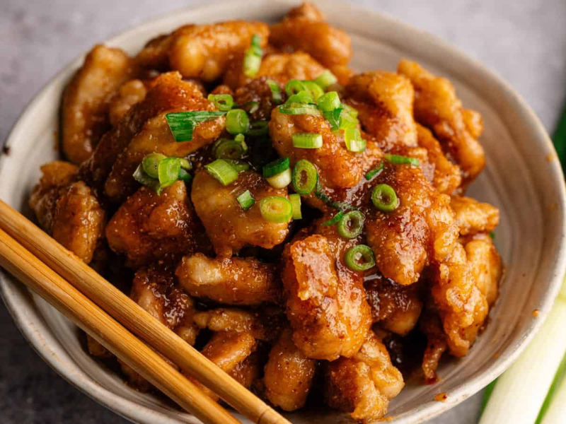 A bowl of glazed chicken pieces topped with chopped green onions, with chopsticks resting on the bowl and green onions beside it.
