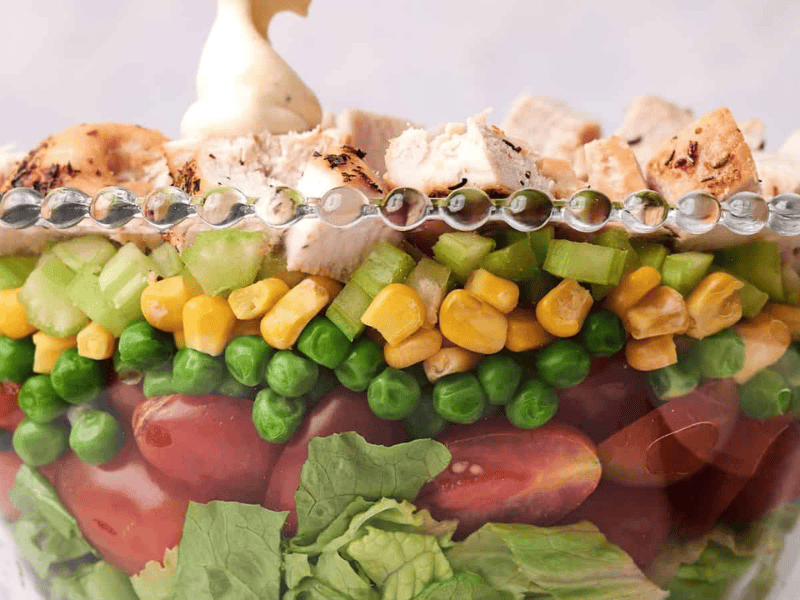 A clear glass bowl layered with lettuce, cherry tomatoes, green peas, corn, diced celery, and topped with chunks of grilled chicken, garnished with a dollop of mayonnaise.
