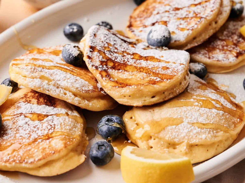 A plate of pancakes topped with powdered sugar, fresh blueberries, lemon wedges, and drizzled with syrup.