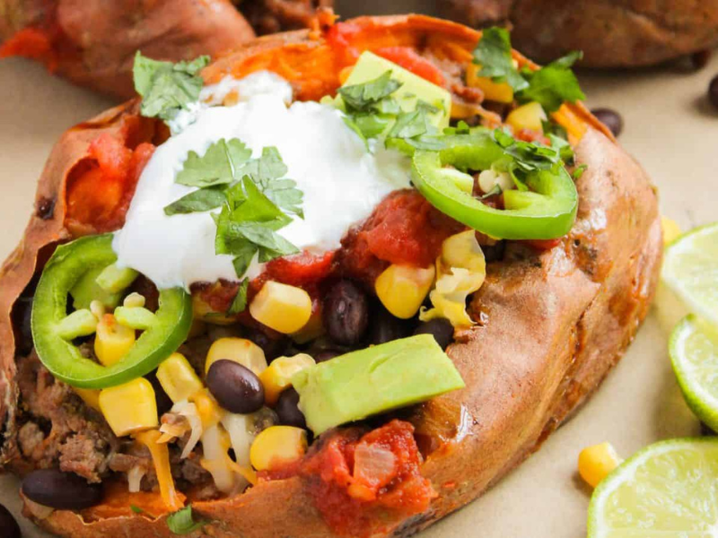 Baked sweet potato topped with black beans, corn, avocado, sliced jalape&ntilde;os, cheese, salsa, sour cream, and cilantro, with lime slices on the side.