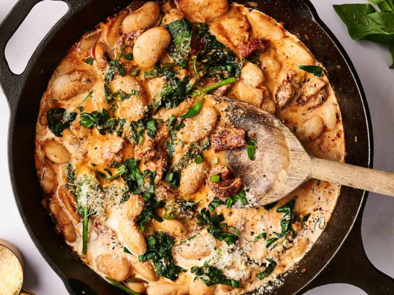 A cast iron skillet with creamy pasta mixed with spinach and sun-dried tomatoes, topped with cheese, with a wooden spoon resting inside.