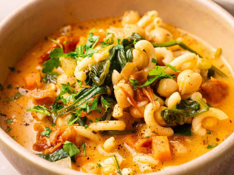 A bowl of pasta soup with chickpeas, spinach, tomatoes, and herbs in a creamy broth.