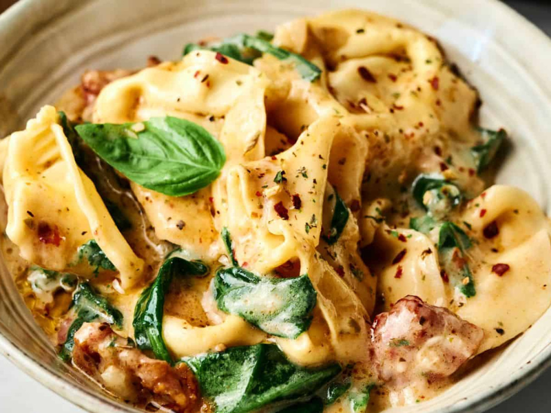 A bowl of creamy tortellini pasta with spinach, sun-dried tomatoes, herbs, and a basil garnish.