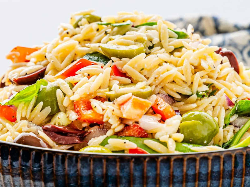 A bowl of orzo salad with mixed vegetables, including olives, tomatoes, and spinach, garnished with herbs.