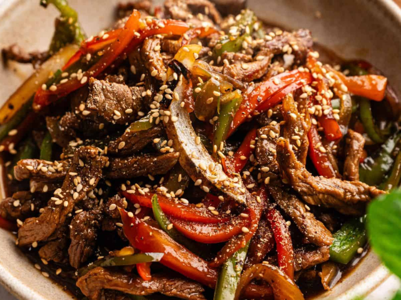 A bowl of stir-fried beef strips with red and green bell peppers, topped with sesame seeds.