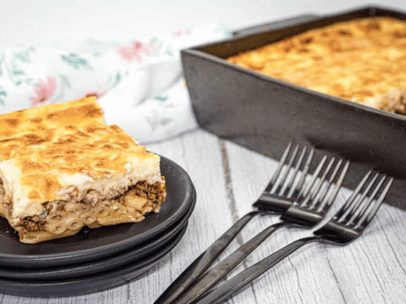 A square piece of pastitsio served on a stack of black plates with three forks and a rectangular baking tray filled with the same pasta on the side.