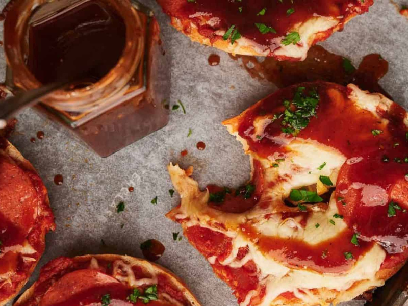 Pizza bagels on parchment paper, one with a bite out of it and a jar of BBQ sauce nearby.