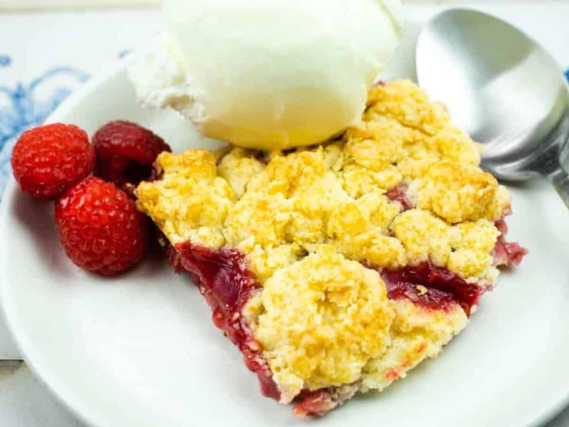 A slice of berry cobbler topped with a scoop of vanilla ice cream is served on a white plate with a spoon and fresh raspberries.