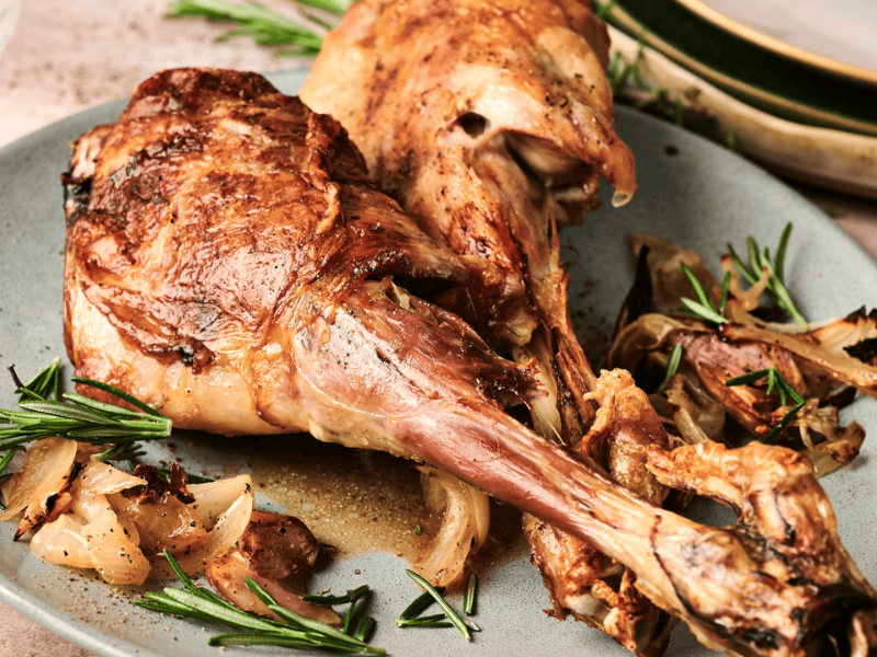 Two roasted lamb shanks on a plate with rosemary and caramelized onions, with stacked plates in the background.