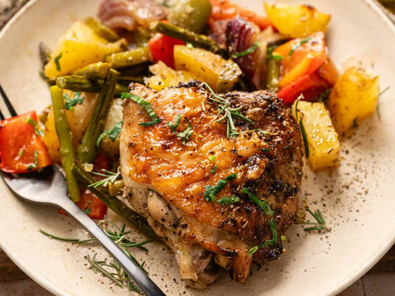A plate with roasted chicken thigh, mixed roasted vegetables including potatoes, green beans, red onions, and bell peppers, garnished with herbs.