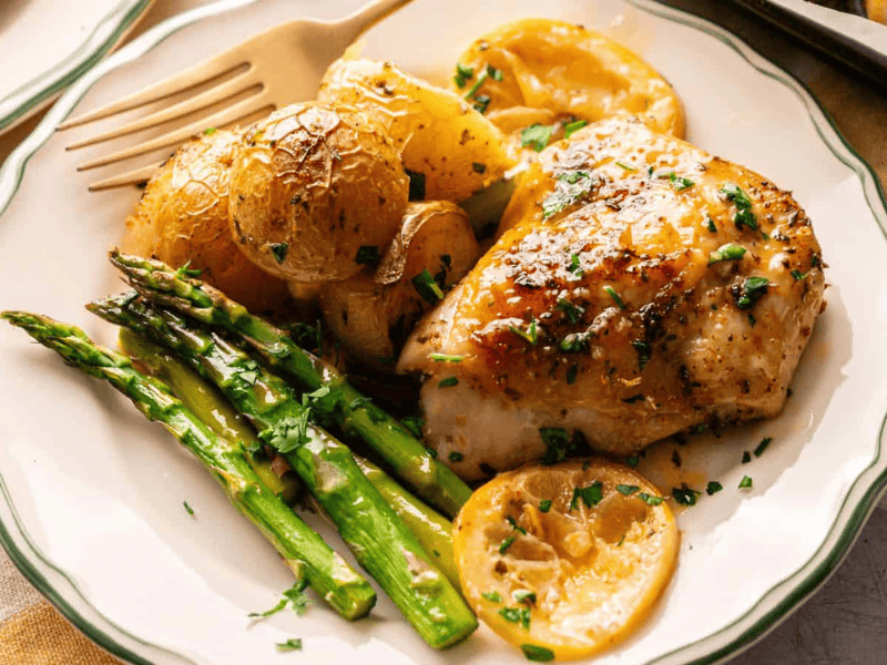 A plate with roasted chicken breast, golden potatoes, asparagus spears, and lemon slices, garnished with chopped herbs.