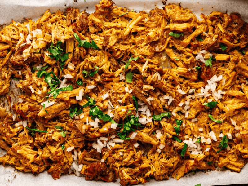 Shredded, seasoned meat spread on a parchment-lined baking sheet, topped with chopped onions and fresh cilantro.