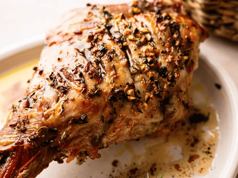 A cooked, herb-crusted leg of lamb rests on a white plate, garnished with seasonings and juices visible on the dish.