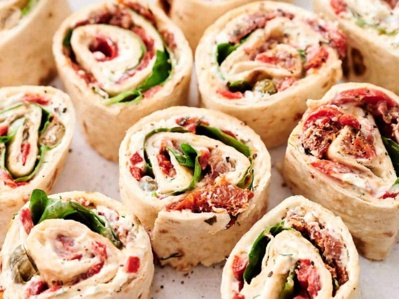 Close-up of several tortilla pinwheel wraps filled with various ingredients including spinach, cream cheese, tomatoes, and other vegetables, arranged neatly on a white surface.