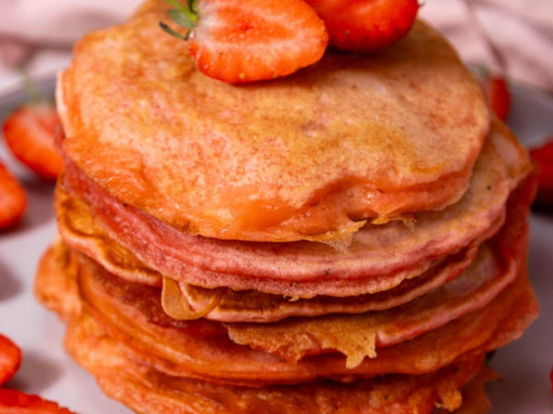 A stack of pancakes topped with sliced strawberries, surrounded by more strawberries on a plate.